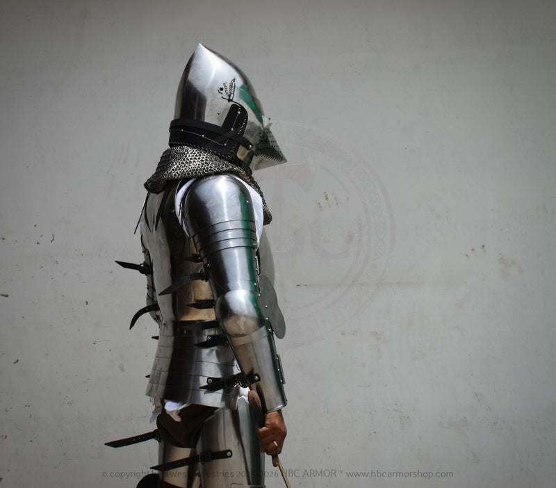 A person wearing medieval-style steel plate armor and a helmet, shown in profile against a plain light gray wall. The armor includes polished steel sections, chainmail neck protection, and dark straps.