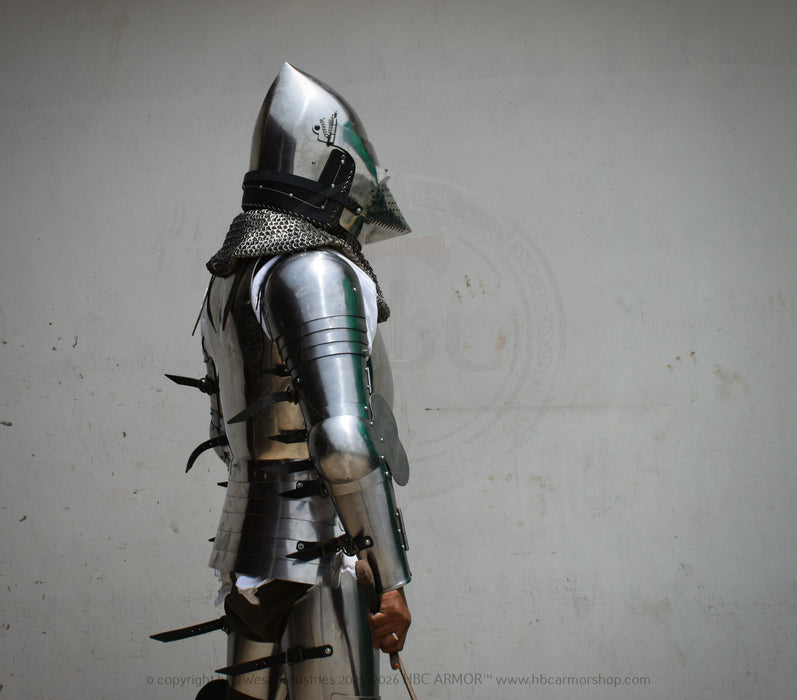 A person wearing medieval-style steel plate armor and a helmet, shown in profile against a plain light gray wall. The armor includes polished steel sections, chainmail neck protection, and dark straps.