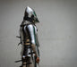 A person wearing medieval-style steel plate armor and a helmet, shown in profile against a plain light gray wall. The armor includes polished steel sections, chainmail neck protection, and dark straps.