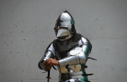 A person wearing full medieval-style steel armor with a polished helmet, chainmail neck guard, and articulated breastplate, standing against a plain light gray wall and holding a sword hilt.