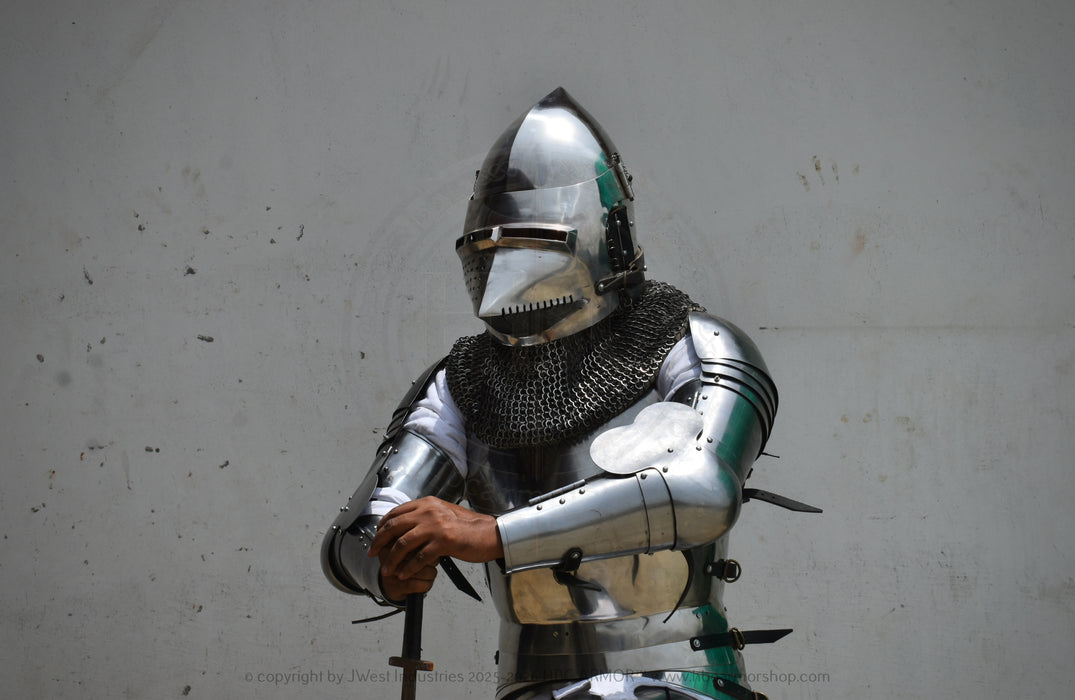 A person wearing full medieval-style steel armor with a polished helmet, chainmail neck guard, and articulated breastplate, standing against a plain light gray wall and holding a sword hilt.