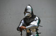A person wearing full medieval-style steel armor with a polished helmet, chainmail neck guard, and articulated breastplate, standing against a plain light gray wall and holding a sword hilt.