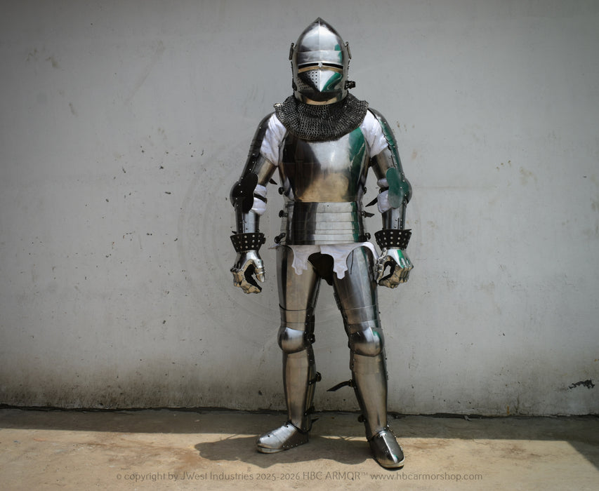 Full-body medieval plate armor suit in polished silver steel with chainmail neck guard, standing against a plain light gray wall.