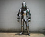 Full-body medieval plate armor suit in polished silver steel with chainmail neck guard, standing against a plain light gray wall.