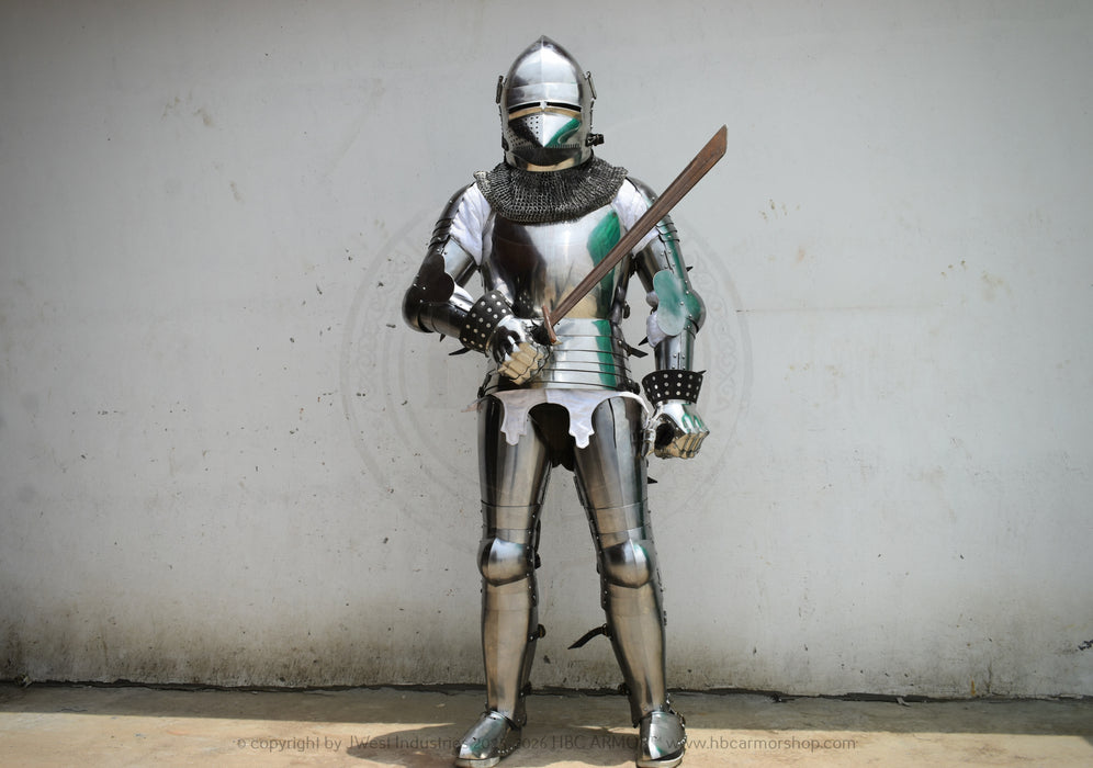 A full suit of medieval plate armor with a helmet and chainmail neck guard, shown standing against a plain light gray wall. The armor has silver steel plates with green painted accents on the chest, shoulders, and lower legs, and the wearer holds a wooden sword.