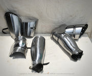 A set of medieval-style steel plate armor pieces, including a breastplate, gauntlets, and shin guards, displayed on a white surface against a white fabric backdrop.