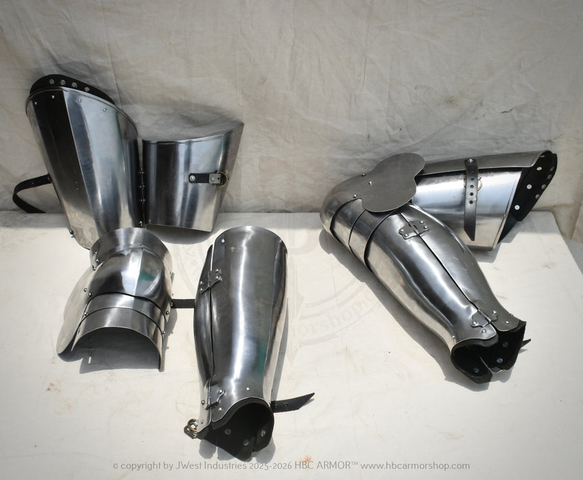 A set of medieval-style steel plate armor pieces, including a breastplate, gauntlets, and shin guards, displayed on a white surface against a white fabric backdrop.