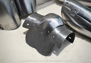 A dark metallic plate armor piece with a curved, multi-panel design, displayed on a light concrete floor alongside other steel armor components.