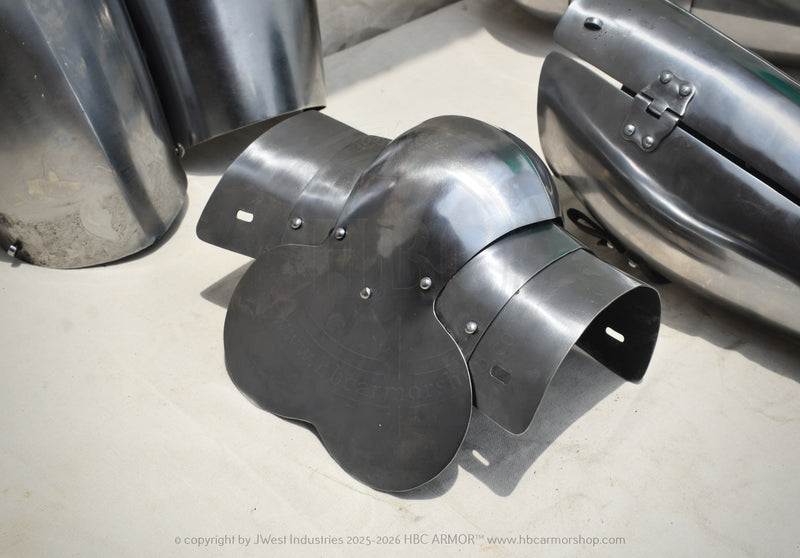 A dark metallic plate armor piece with a curved, multi-panel design, displayed on a light concrete floor alongside other steel armor components.