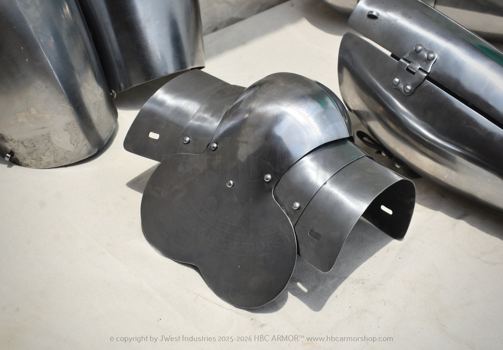 A dark metallic plate armor piece with a curved, multi-panel design, displayed on a light concrete floor alongside other steel armor components.